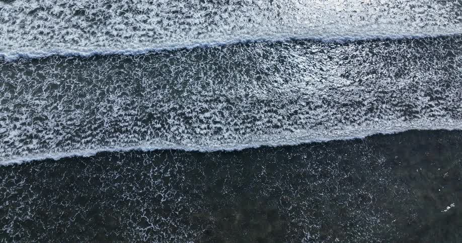 A mesmerizing top-down aerial view captures the textured patterns of dark ocean waves rolling and breaking in the sunlight
