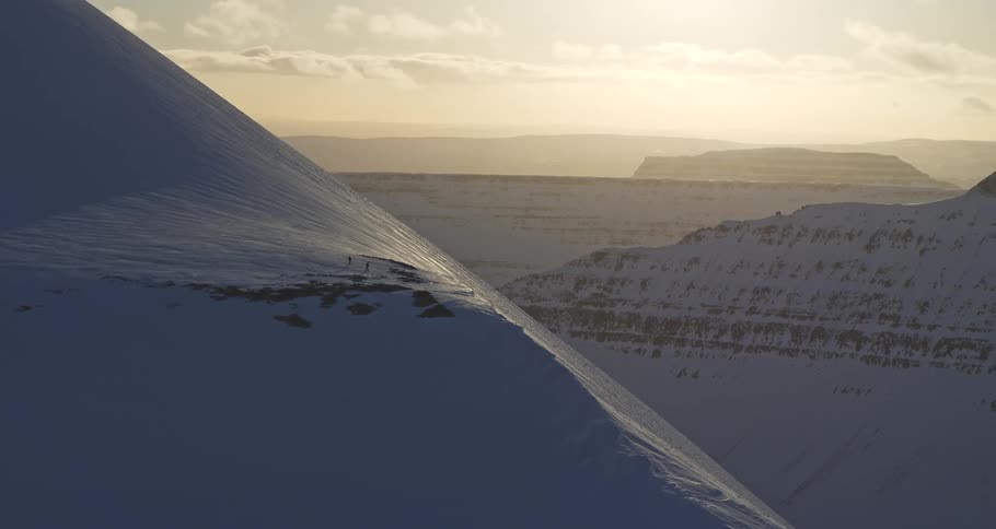 An epic aerial shot reveals two distant skiers standing on a vast snowy mountain ridge as the sun sets over the mountain range