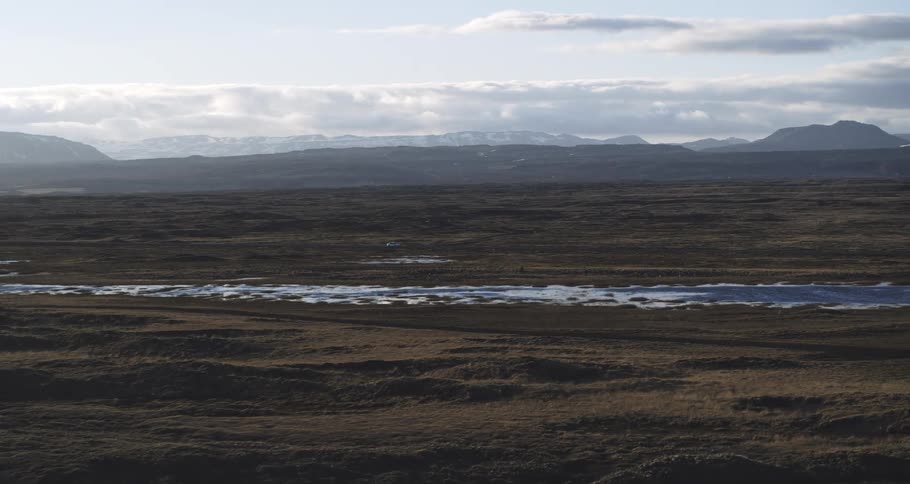 An aerial shot reveals a lone white car driving through a vast, moody Icelandic landscape with snow-capped mountains in the distance
