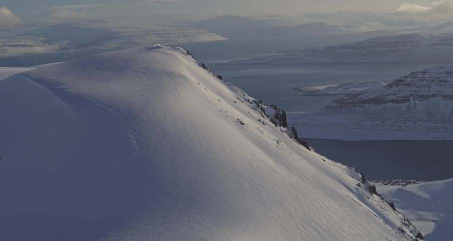 A breathtaking high-angle view of a pristine, snow-covered mountain ridge overlooking a vast fjord system under a winter sky