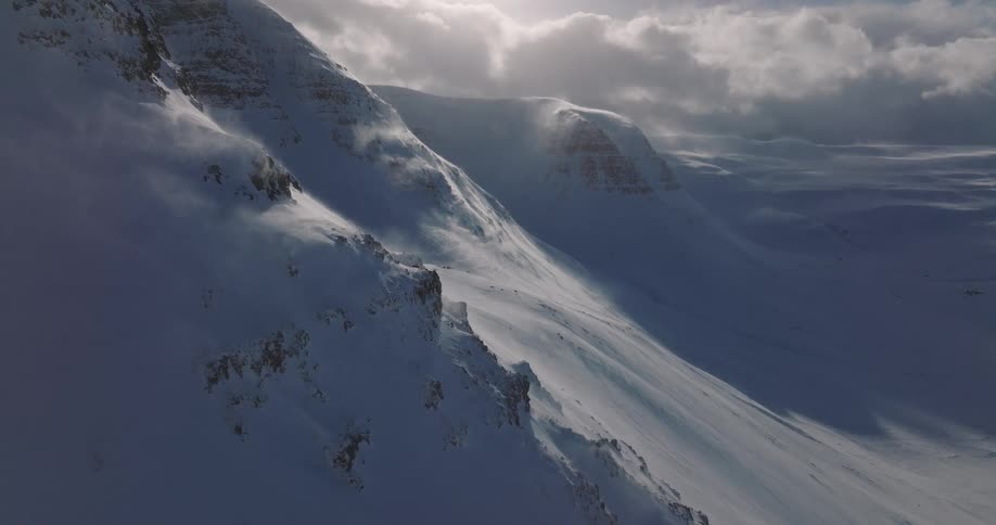 An epic aerial view shows strong winds blowing snow off the peaks and ridges of a majestic, desolate mountain landscape