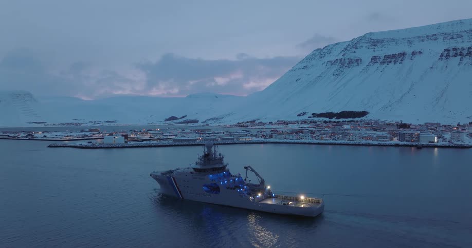 A majestic Icelandic Coast Guard vessel sails through a snowy fjord at twilight, with the lights of a coastal town gleaming in the background