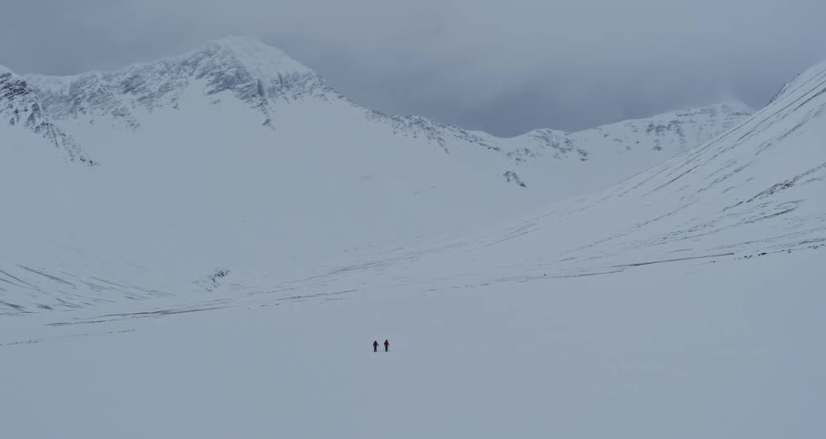 An epic aerial view follows two explorers walking through a vast, remote, and snow-covered mountain valley