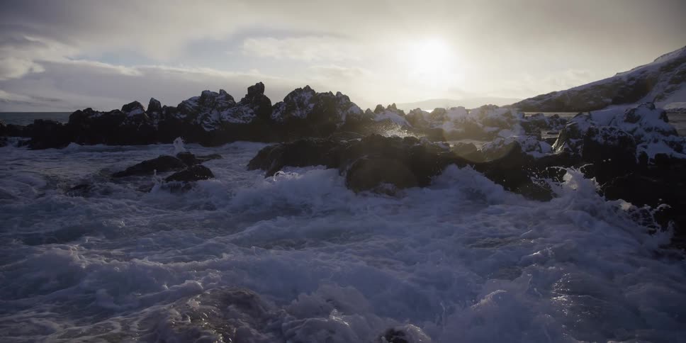 Powerful ocean waves crash against a snow-covered rocky coastline in dramatic slow motion at sunrise