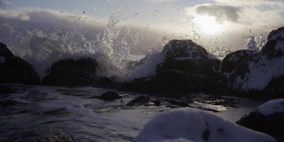 A powerful ocean wave crashes in slow motion against snow-dusted rocks on a wintery shoreline at sunset