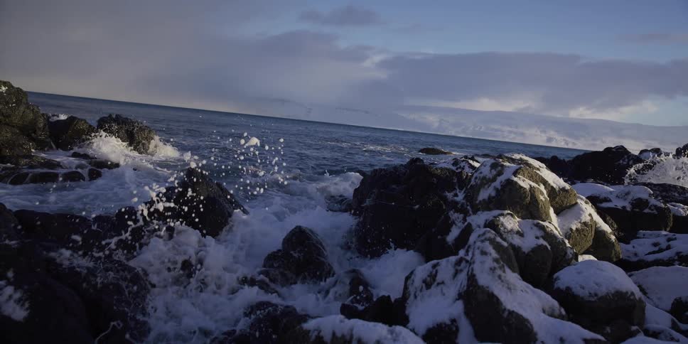 Icy ocean waves crash in dramatic slow motion against a shoreline of snow-covered rocks with mountains in the distance