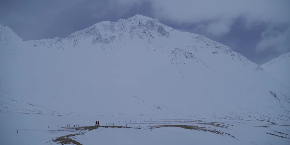 Two hikers are dwarfed by the immense scale of a snow-covered mountain as they trek across a vast, desolate winter landscape