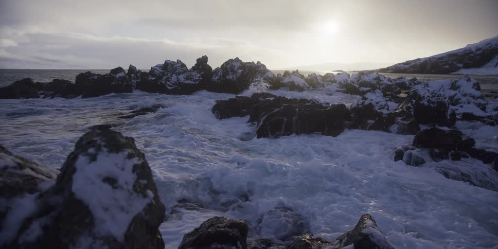 Dramatic slow-motion footage shows powerful ocean waves crashing against a snowy and rocky coastline under a bright, overcast sun