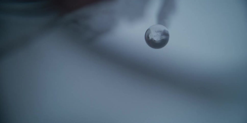 An extreme close-up shot captures the slow process of a water droplet forming and growing at the tip of a melting icicle