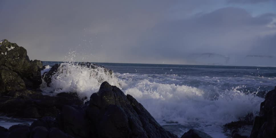 Powerful ocean waves crash against a dark rocky shoreline in dramatic slow motion
