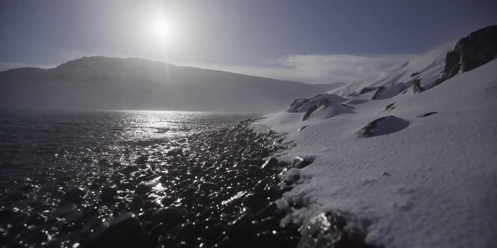 A slow dolly shot reveals a rocky, snow-covered beach on the edge of a frigid sea under the bright sun
