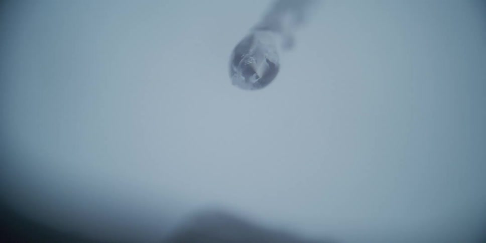 A slow-motion macro shot captures a single drop of water forming on the tip of a melting icicle before falling