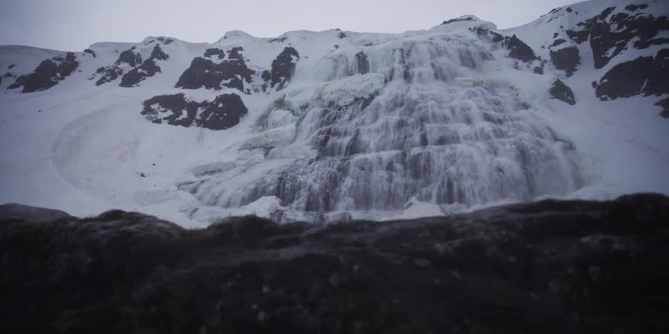 A massive waterfall flows down a snow-covered mountain in a cold, dramatic landscape
