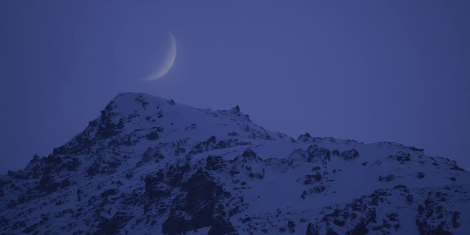A crescent moon hangs in the deep blue twilight sky above a snowy mountain peak