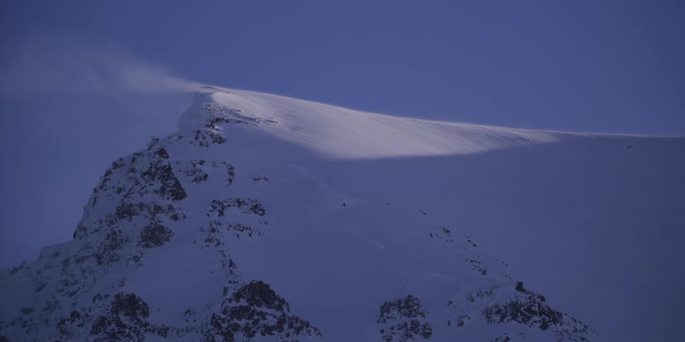 Strong winds blow snow across a majestic, sun-kissed mountain peak at dusk
