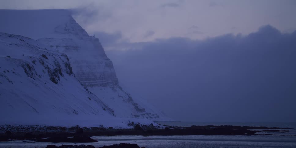 A moody, tranquil shot of a snow-covered mountain meeting the cold sea at dusk in Iceland
