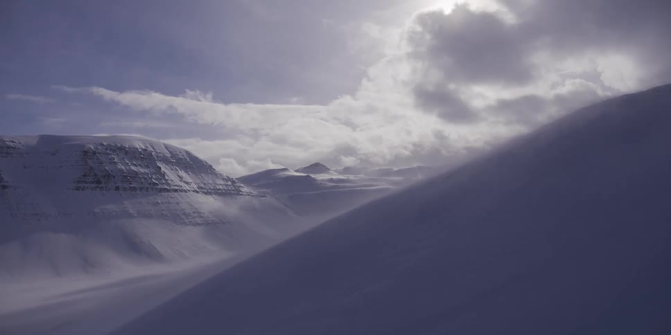 Strong winds whip snow across a vast, frozen mountain landscape under a moody, sunlit sky
