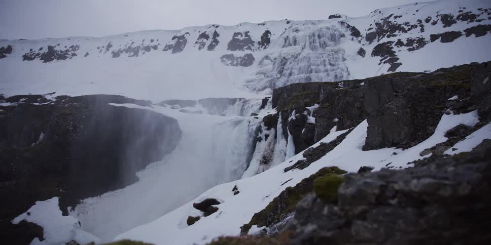 A majestic, partially frozen waterfall cascades down a snowy mountain cliff on a cold, overcast day