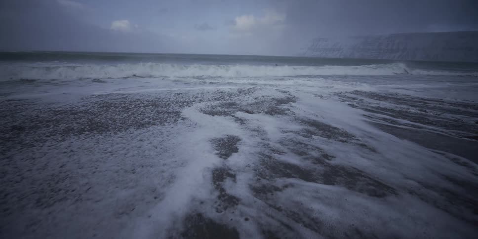 Slow-motion waves retreat over a dark sand beach with snowy mountains in the distance on a cold, overcast day