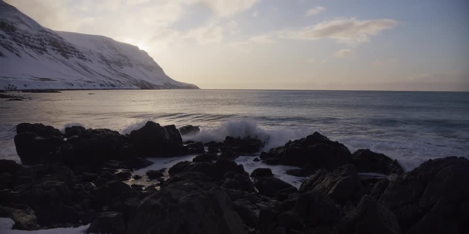 Slow motion footage shows ocean waves crashing against a rocky shore with a snow-covered mountain in the background