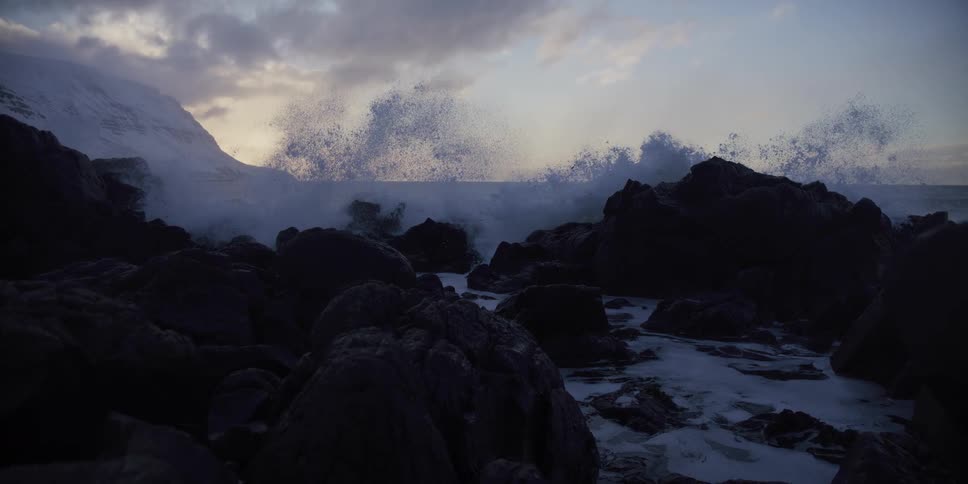 A powerful ocean wave crashes in dramatic slow motion against a rugged, snowy coastline at dusk