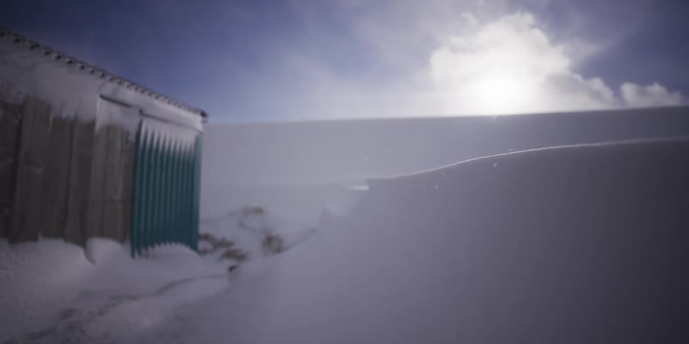 A slow-motion dolly shot captures blowing snow sweeping across a pristine drift next to a weathered building