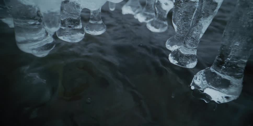 A mysterious close-up of dark water flowing beneath bulbous icicles in a winter stream