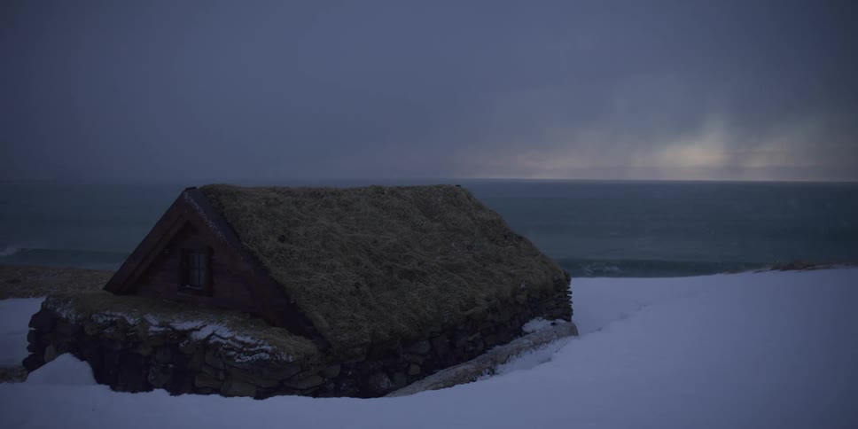 A traditional turf-roofed stone cabin stands on a snowy coastline as a winter storm blows in from the sea