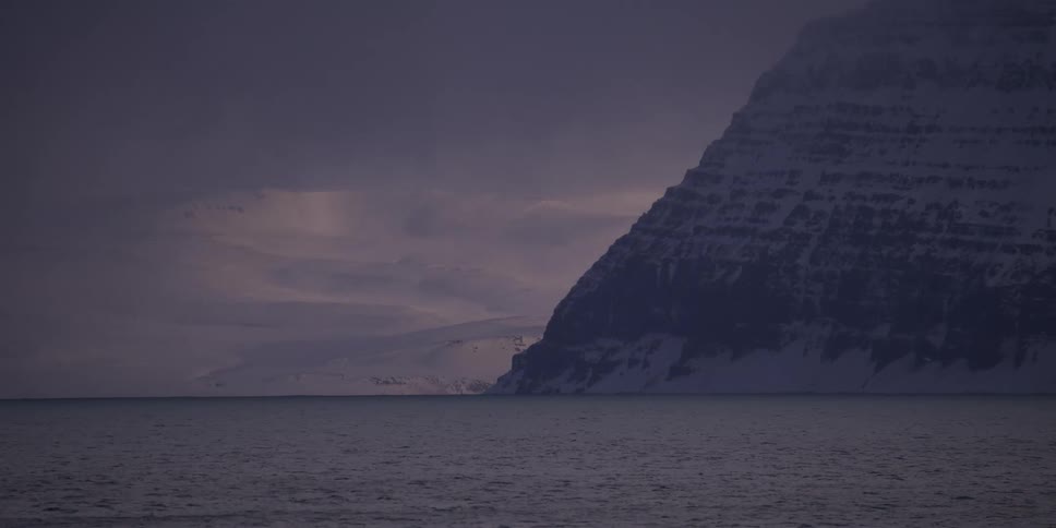 A dramatic, snow-covered mountain range looms over a dark, tranquil sea under a heavy, overcast winter sky