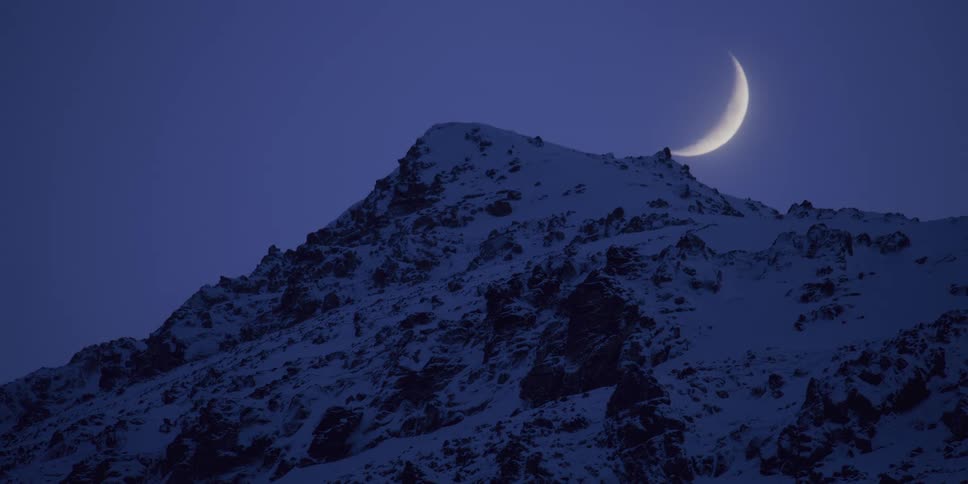 A bright crescent moon appears to rest on the peak of a snow-covered mountain against a dark blue night sky