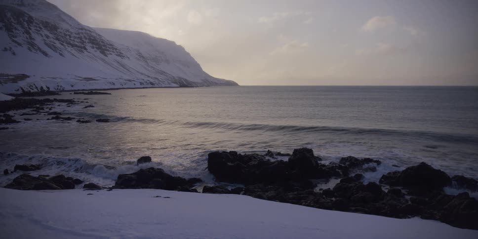Calm ocean waves lap against a snowy and rocky shore below a majestic, snow-covered mountain at dusk