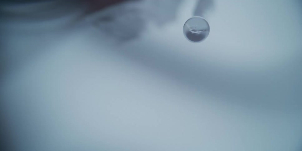 A macro slow-motion shot captures a clear liquid droplet forming and detaching from the tip of a pipette