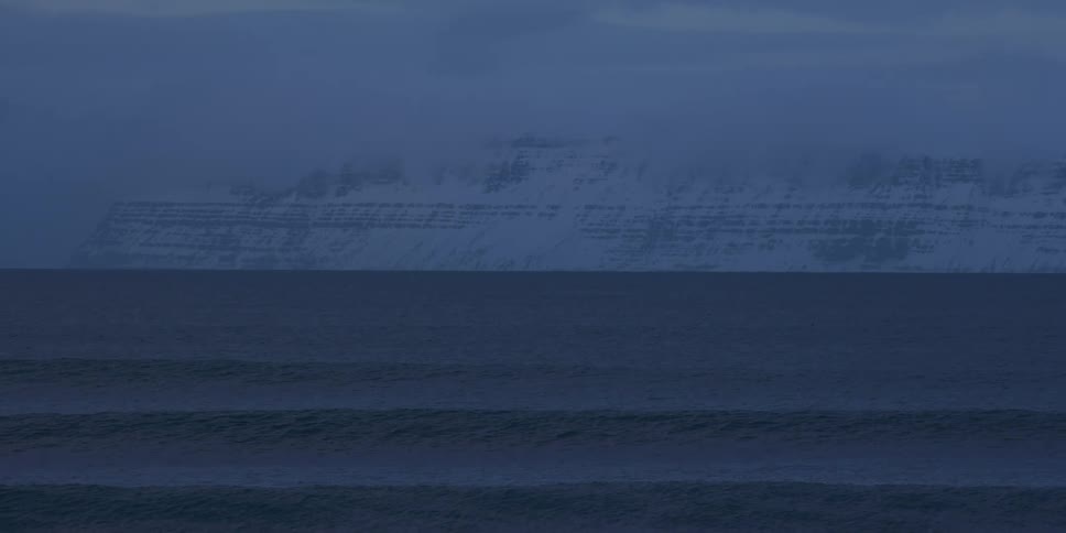 Gentle ocean waves roll towards the shore with a majestic, snow-covered mountain in the distance at dusk