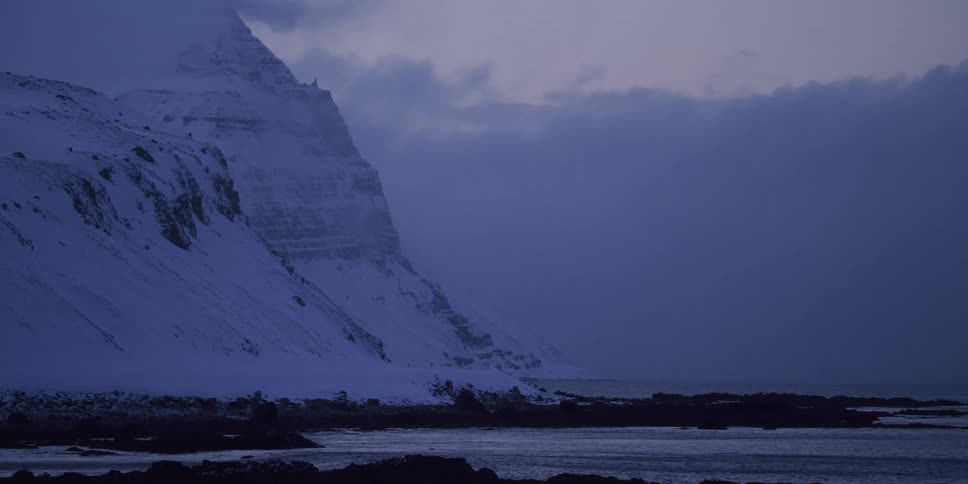 A moody, blue hour view of a snow-covered mountain range meeting the rocky coastline of the arctic sea