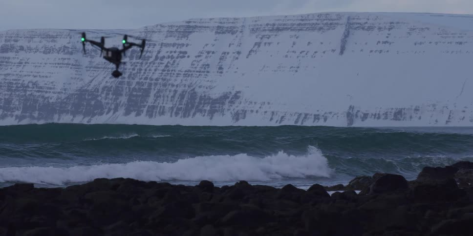 A camera drone hovers in slow motion over a rocky coastline as ocean waves break against the shore with a snowy mountain in the background