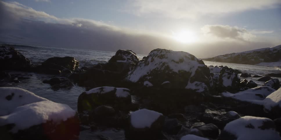 Snow falls in slow motion on a rocky coastline during a cold winter sunset with gentle waves