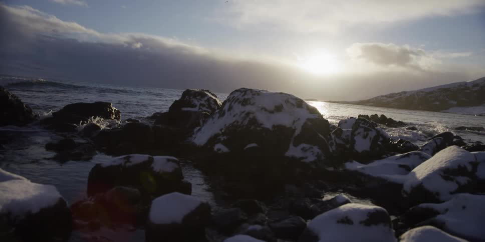 A cinematic slow-motion shot reveals a snowy, rocky shoreline as the sun hangs low over the cold ocean