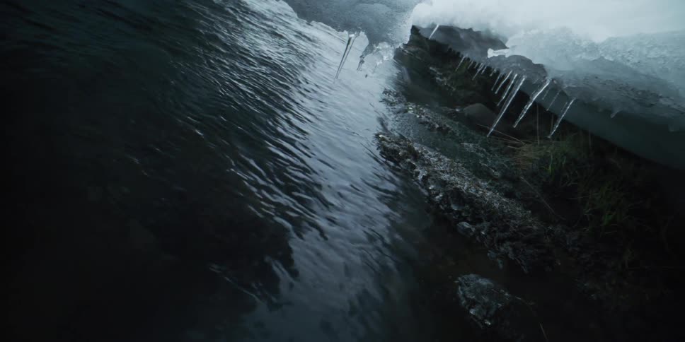 A dark river flows slowly beside a snow-covered bank where sharp icicles hang just above the water's surface