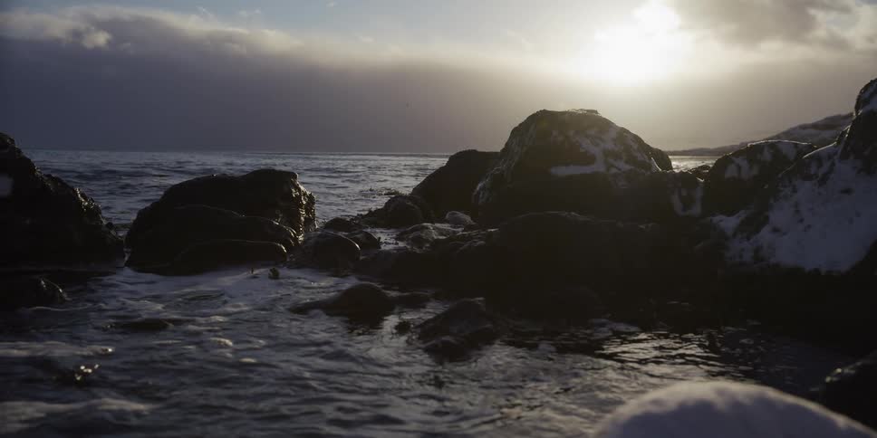 A slow tracking shot reveals a serene winter coastline with snow-capped rocks as the sun hangs low over the ocean
