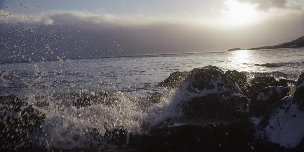 Slow motion waves crash over snow-dusted rocks on a cold shoreline against the setting sun