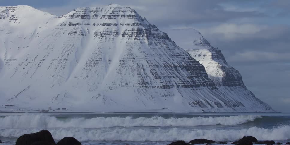 Slow motion shot of ocean waves crashing on a rocky shore with a dramatic snow-covered mountain range in the background