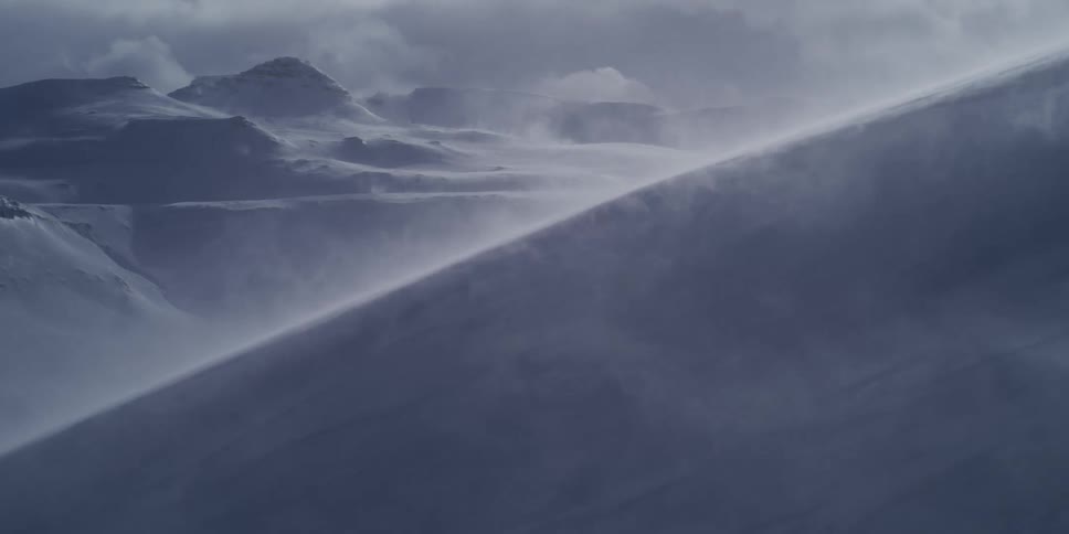 Close up of wind blowing snow across a mountain ridge, revealing a vast and stormy arctic landscape