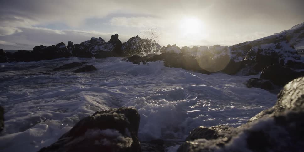 In slow motion, powerful ocean waves crash against an icy, rocky shoreline illuminated by the bright sun
