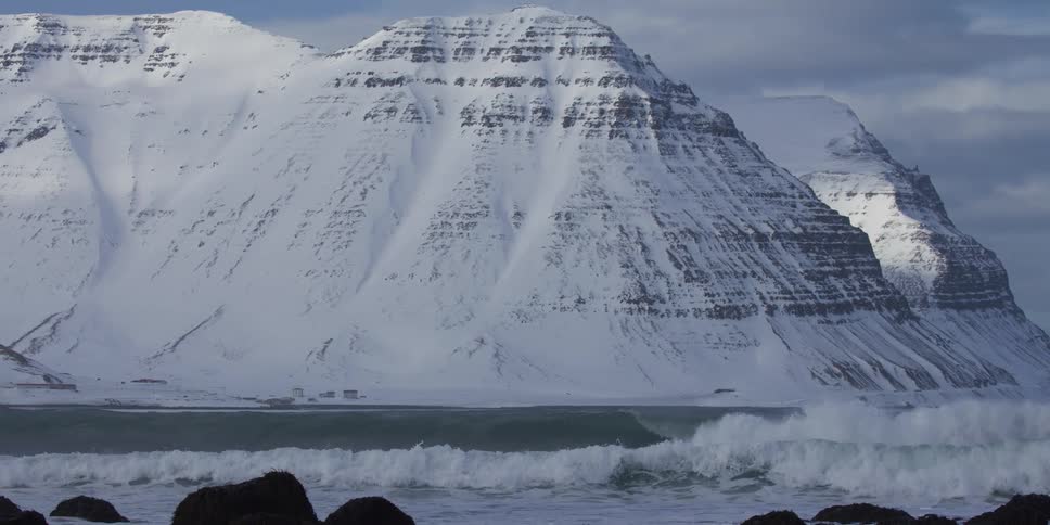 Slow motion waves break along the shore with majestic, snow-covered mountains towering in the background in the Westfjords of Iceland