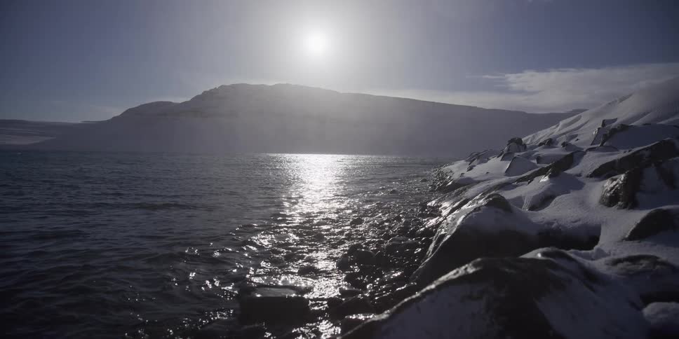 A serene arctic landscape features the sun glittering on the water next to snow-covered mountains and shoreline