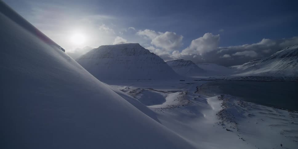 The bright sun shines over a vast, snow-covered mountain landscape next to a dark fjord in the Arctic