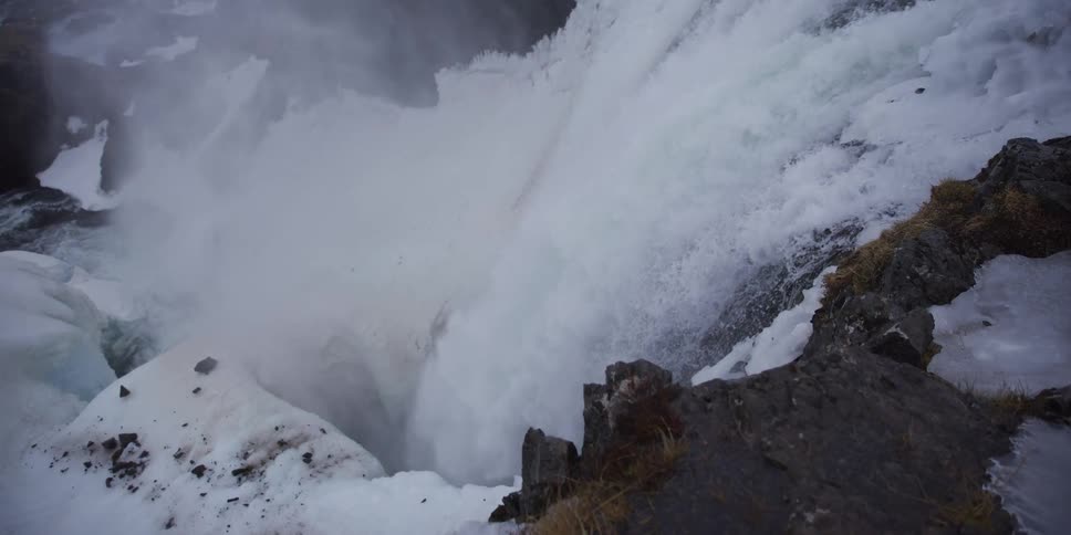 A dramatic, slow-motion shot from the edge of a snowy cliff reveals a powerful waterfall crashing into the icy chasm below