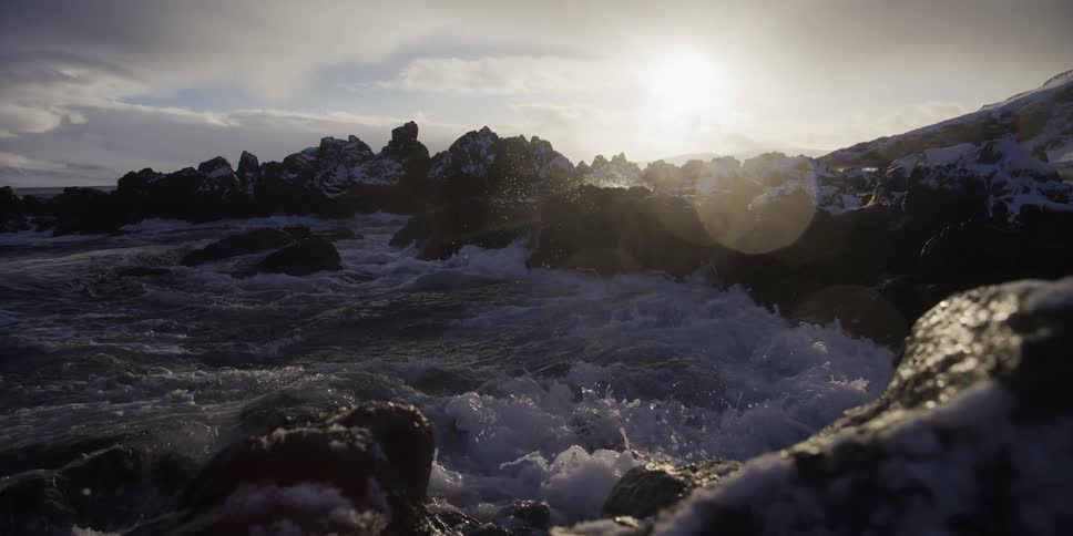 Dramatic slow motion of ocean waves crashing against a snowy, rocky coastline during a beautiful sunset