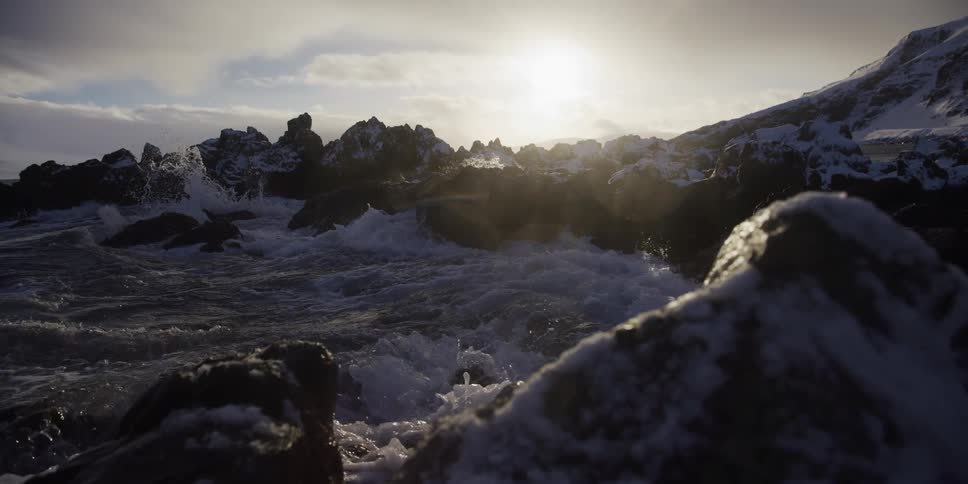 Dramatic slow motion shot of frigid ocean water splashing over snowy volcanic rocks with a bright sun in the background