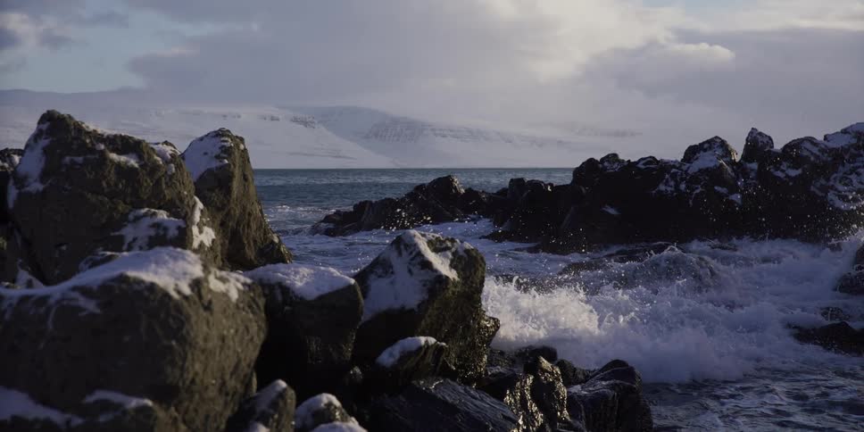 Ocean waves crash in slow motion against a snow-covered rocky coastline with majestic mountains in the distance